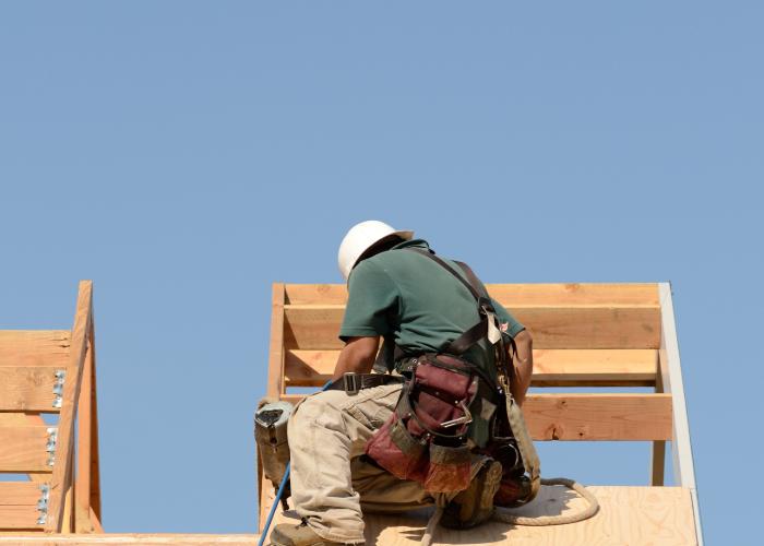 A construction worker, wearing a white hard hat and a green shirt, is seated on a wooden structure in Cranston