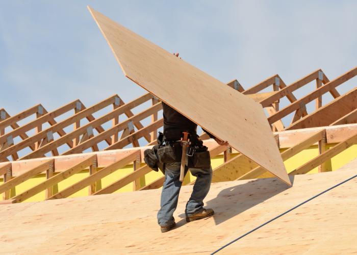 A construction worker carries a large plywood sheet on a roof under construction,