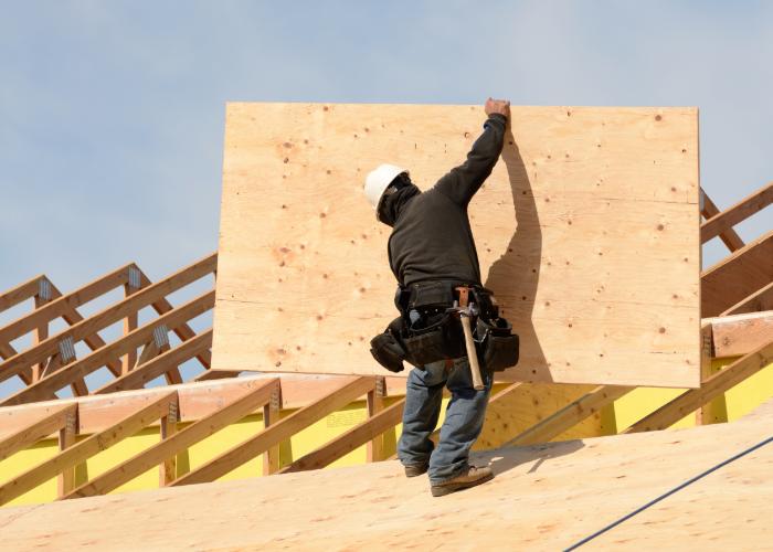 A construction worker in a hard hat installs a large plywood sheet on a roof structure.